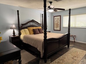 A four-poster bed with a ceiling fan, a nightstand with a lamp, a framed botanical print on the wall, and a window with blinds. - Charming 3/2 house in Jensen Beach with pool and minutes away from downtown (Jensen Beach)