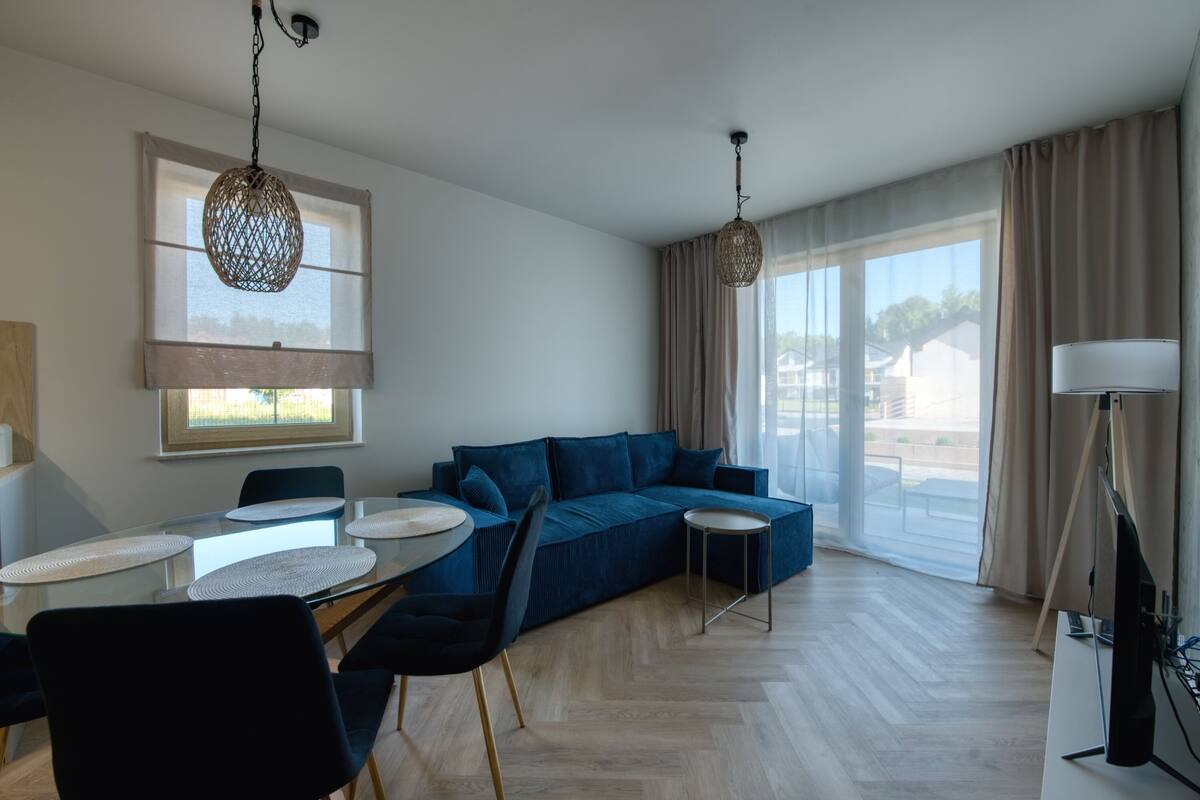 Ein modernes Wohnzimmer mit einer Couch, einem Esstisch mit Stühlen, einem Fernseher und großen Fenstern mit Vorhängen.