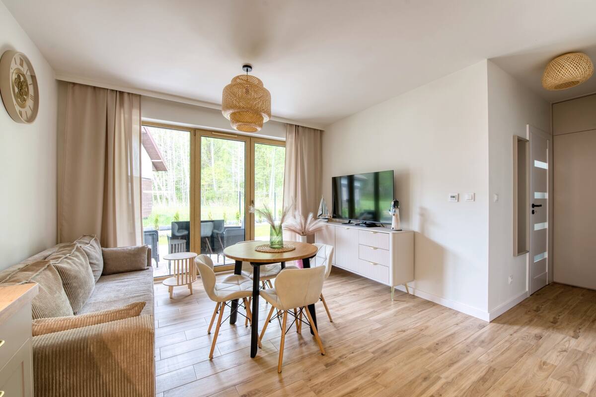 Ein modernes Wohnzimmer mit einer Couch, einem Esstisch, Stühlen, einem Fernseher und einem großen Fenster mit Vorhängen.