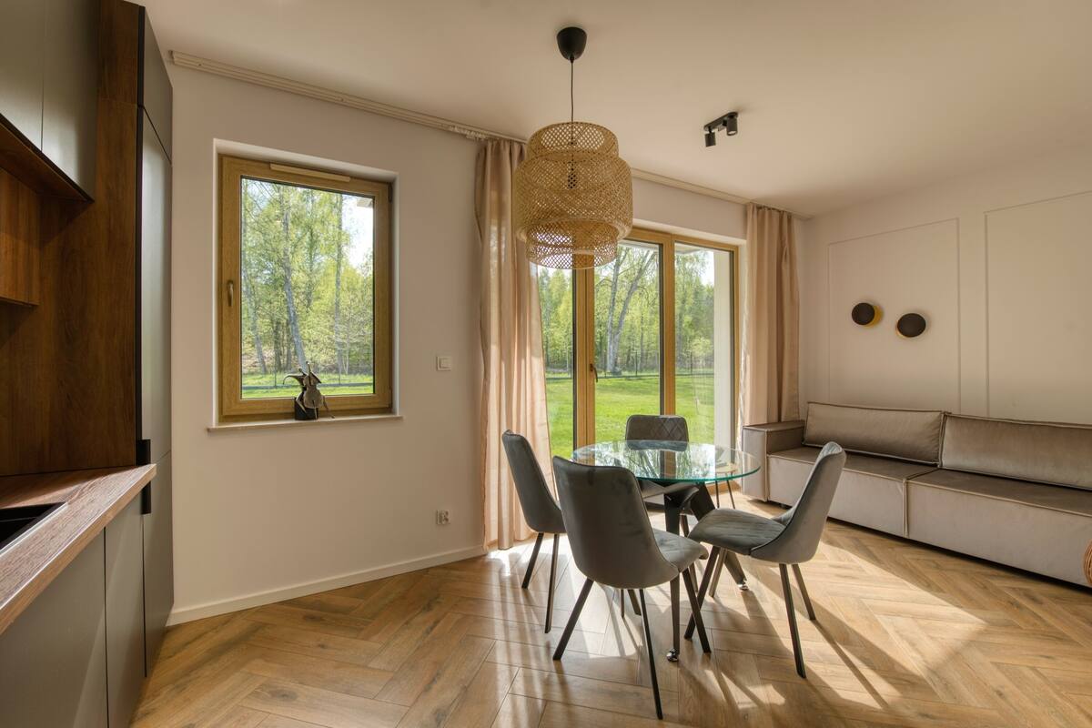 Ein moderner Essbereich mit einem Glastisch, grauen Stühlen, einer gewebten Pendelleuchte und Blick auf eine grüne Landschaft durch große Fenster.