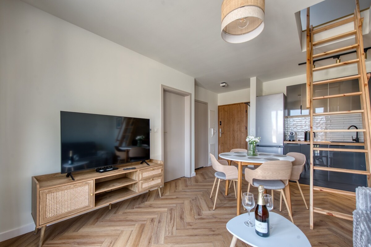 A modern living room with a flat-screen TV, wooden entertainment unit, herringbone-patterned flooring, a dining area with a round table and chairs, and a kitchen with white cabinets and a wooden ladder.