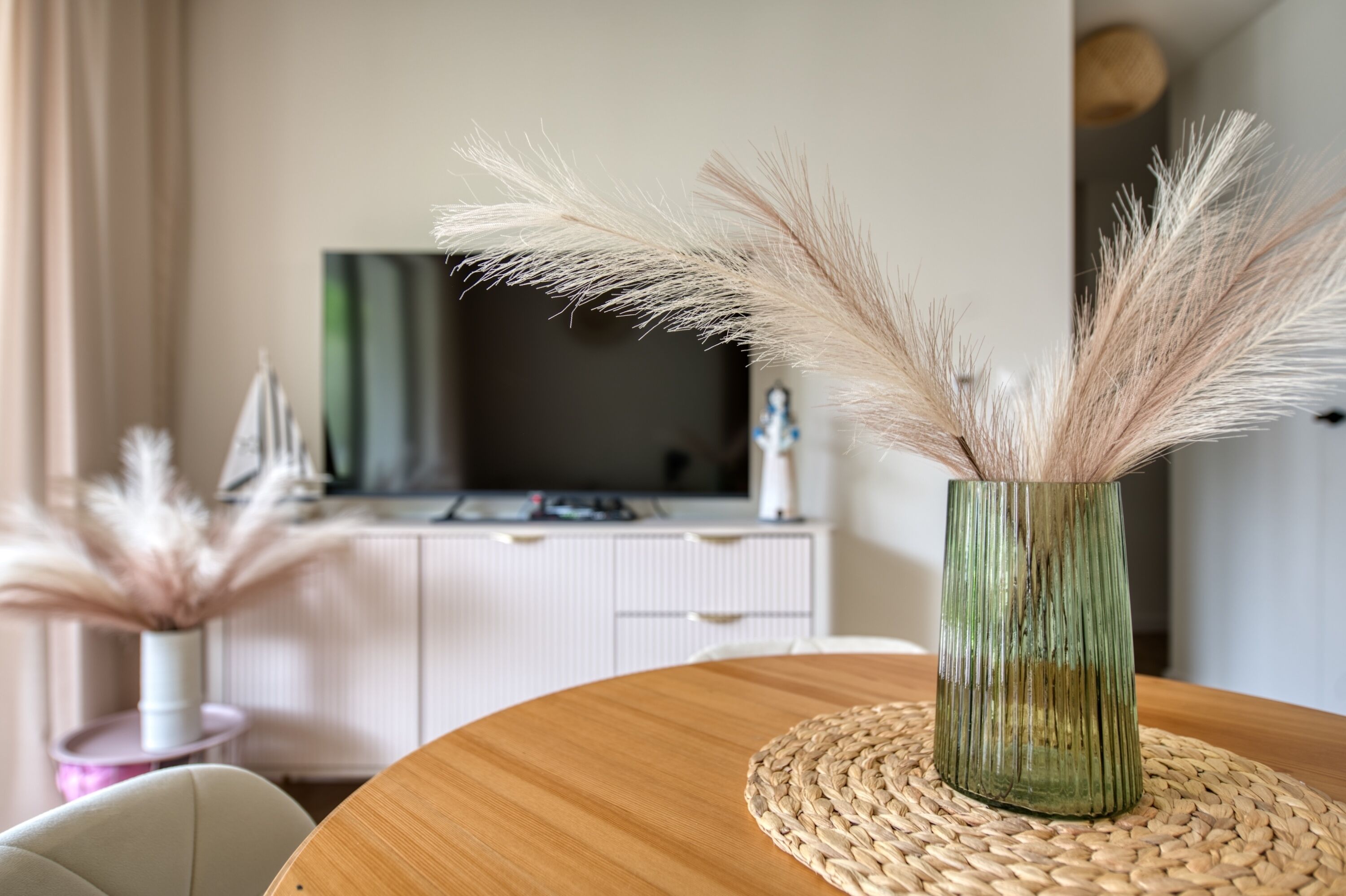Un comedor con una mesa redonda de madera, un mantel individual tejido y un jarrón verde con plumas de pampa.