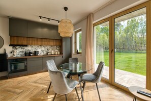 A modern kitchen with a glass dining table and chairs, a large window with a view of a grassy area, and a hanging light fixture.
