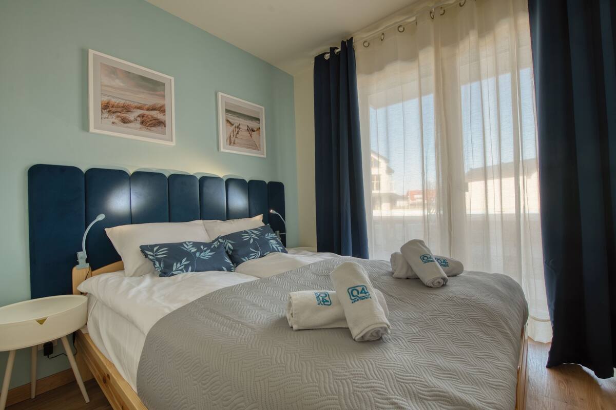 Ein ordentlich bezogenes Bett mit Kopfteil, zwei gerahmte Bilder an der Wand, ein Nachttisch und ein Fenster mit Vorhängen.