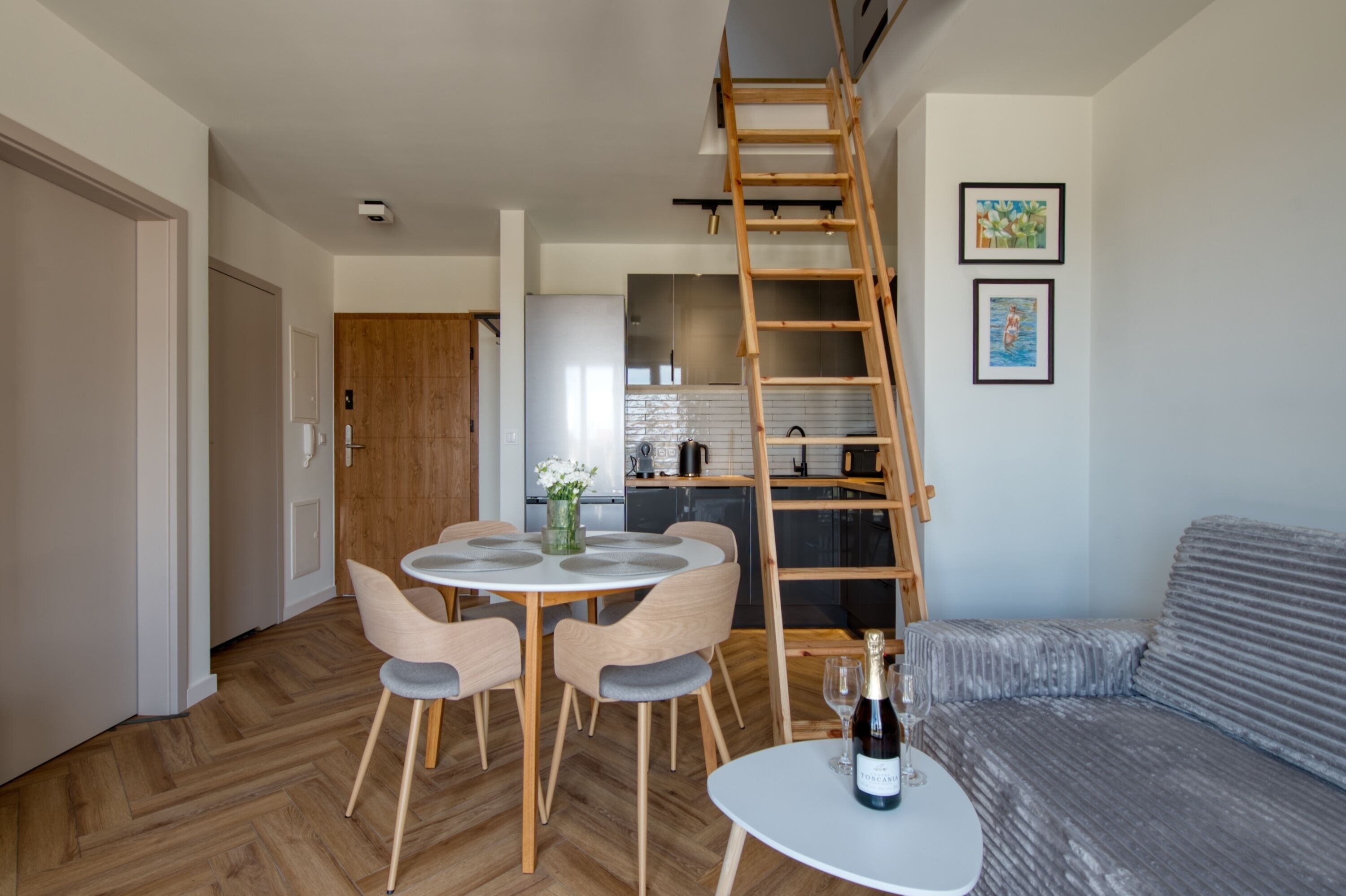 A modern living room with a dining area, a loft bedroom with a wooden ladder, and a kitchen in the background.