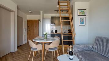 A modern living room with a dining area, a loft bedroom with a wooden ladder, and a kitchen in the background.