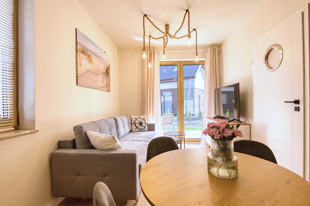 A modern living room with a sofa, a round dining table with a vase of flowers, a television, and a large window with a view of the outside.