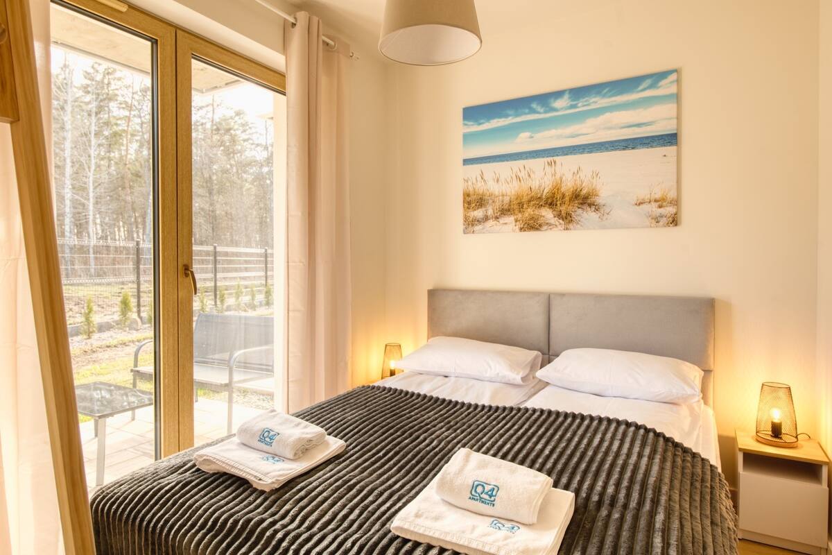 Ein Schlafzimmer mit einem Bett, einem Strandbild, einem Fenster und einem Nachttisch mit einer Lampe.
