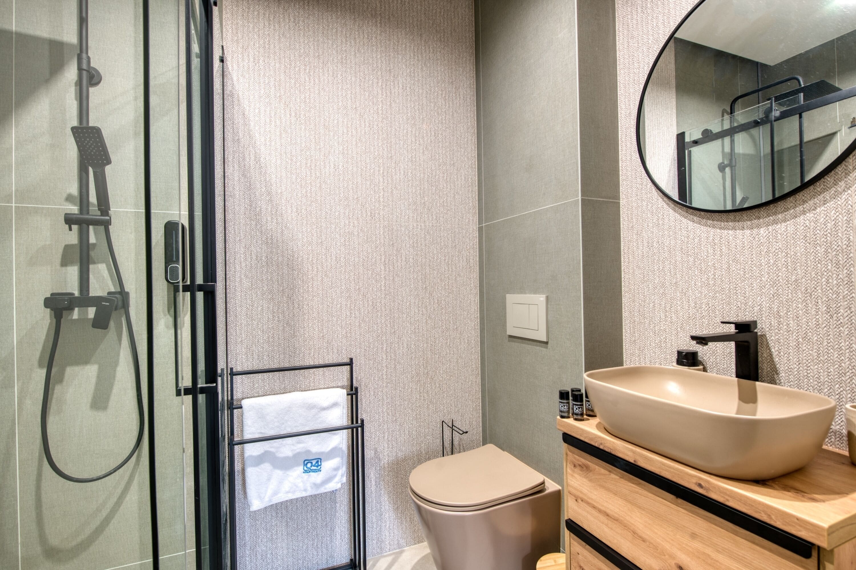 A modern bathroom with a glass-enclosed shower, a beige toilet, a wooden vanity with a large sink, and a round mirror.