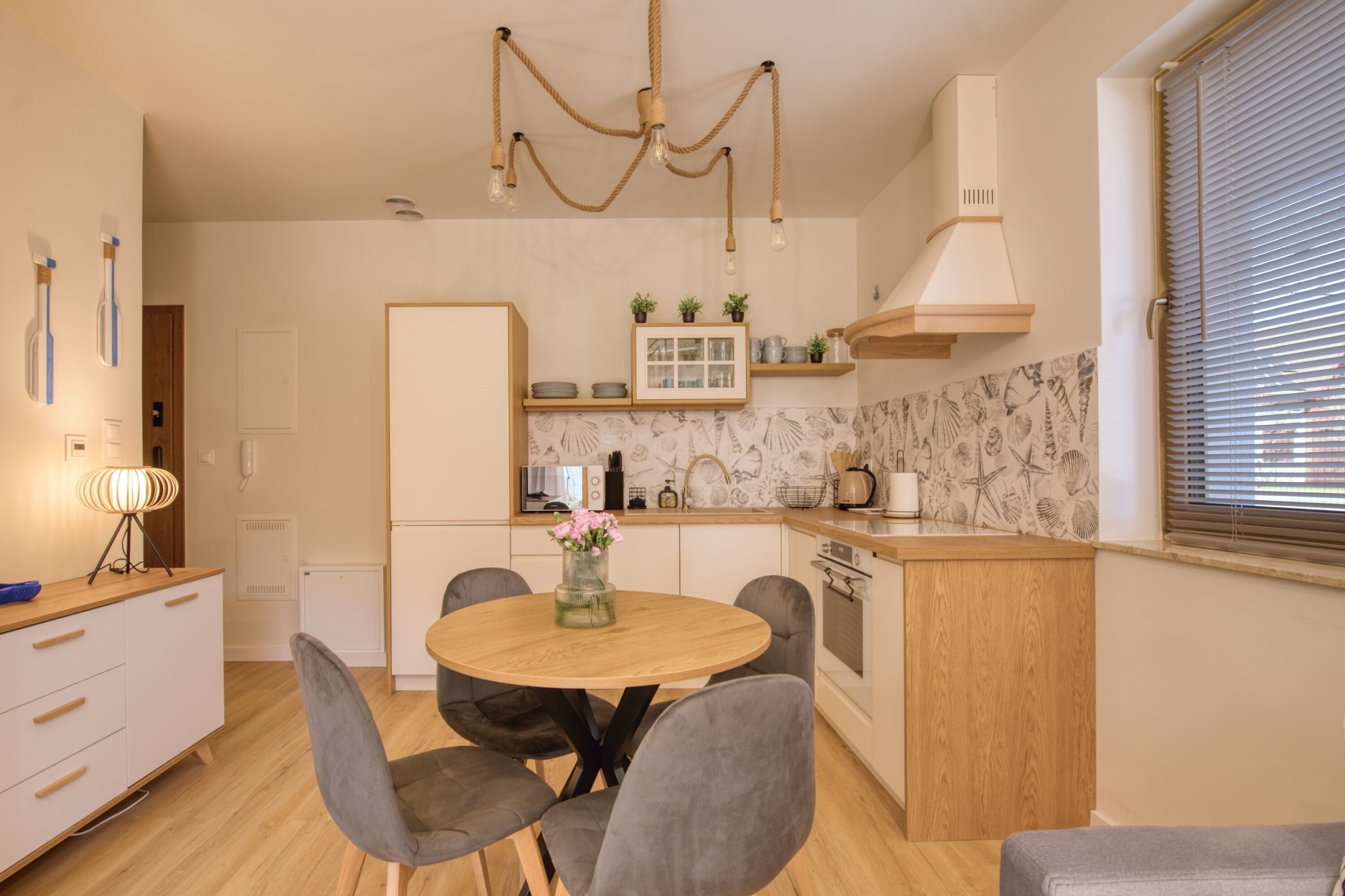 A modern kitchen with a round dining table, grey chairs, a hanging light fixture, and a built-in oven.