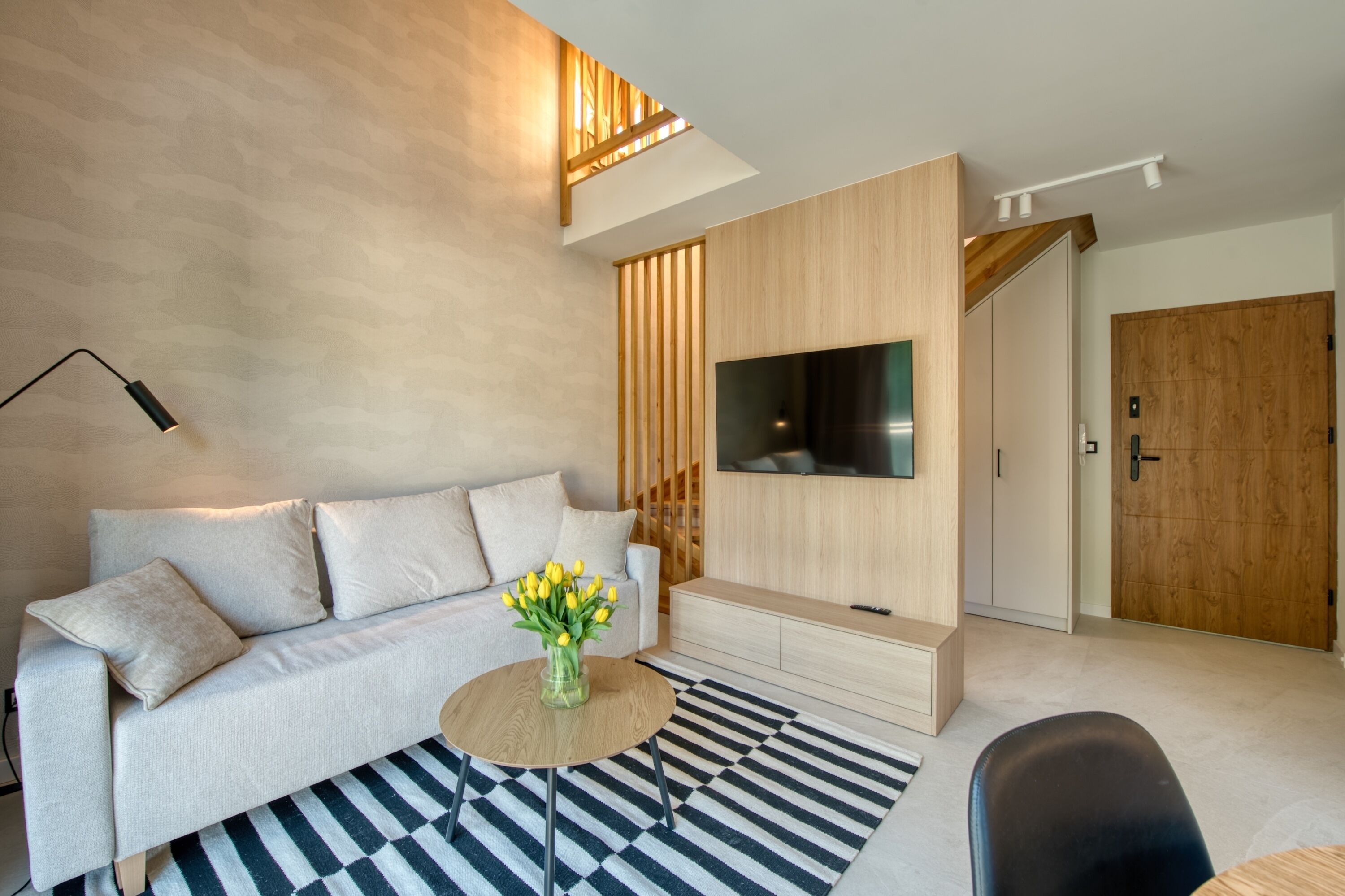 A modern living room with a sofa, a coffee table with flowers, a flat-screen TV, and a wooden door.