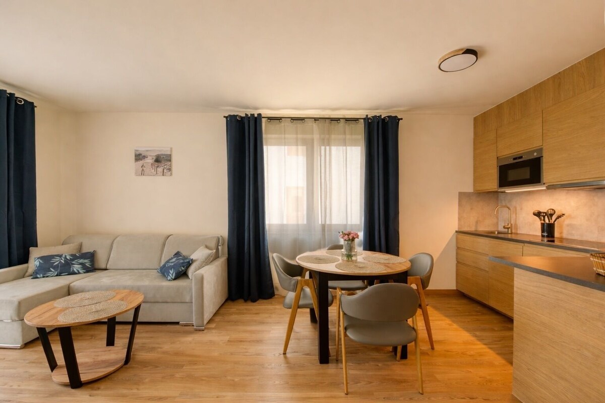 A modern living room with a sofa, a round dining table with chairs, a kitchen area with wooden cabinets and a microwave, and a large window with curtains.