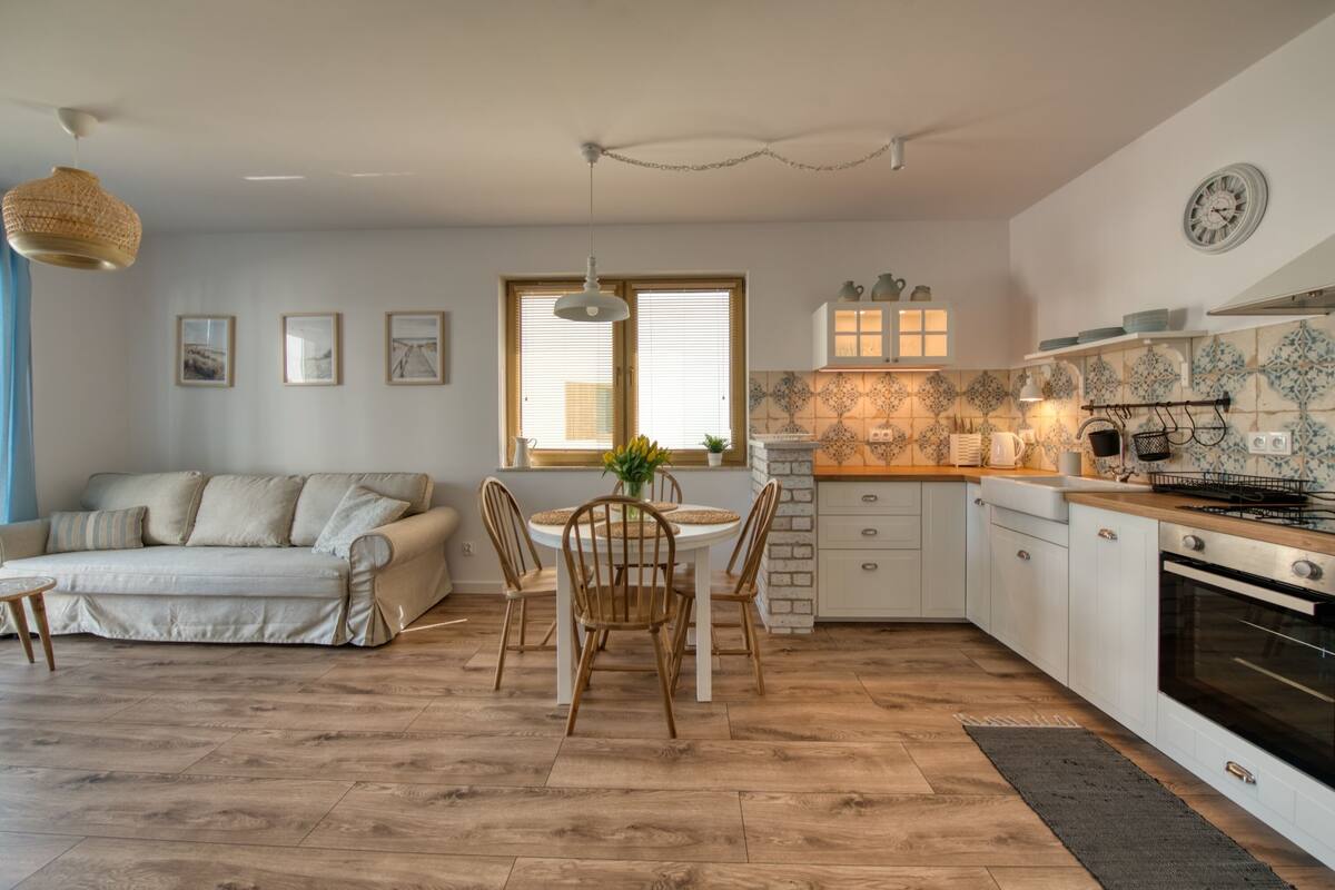 Eine moderne Küche mit weißen Schränken, eine Essecke mit rundem Tisch und Stühlen sowie ein Wohnbereich mit einer Couch und einer Pendelleuchte.