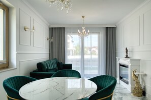 Una habitación con una mesa de mármol, sillas tapizadas en verde, un candelabro, una chimenea con una estatua de pavo real, y vistas a través de amplios ventanales.