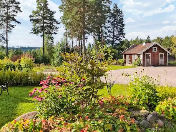 Uma cabana rústica com entrada por um caminho de pedras e um jardim bem cuidado, com diversas plantas e flores.
