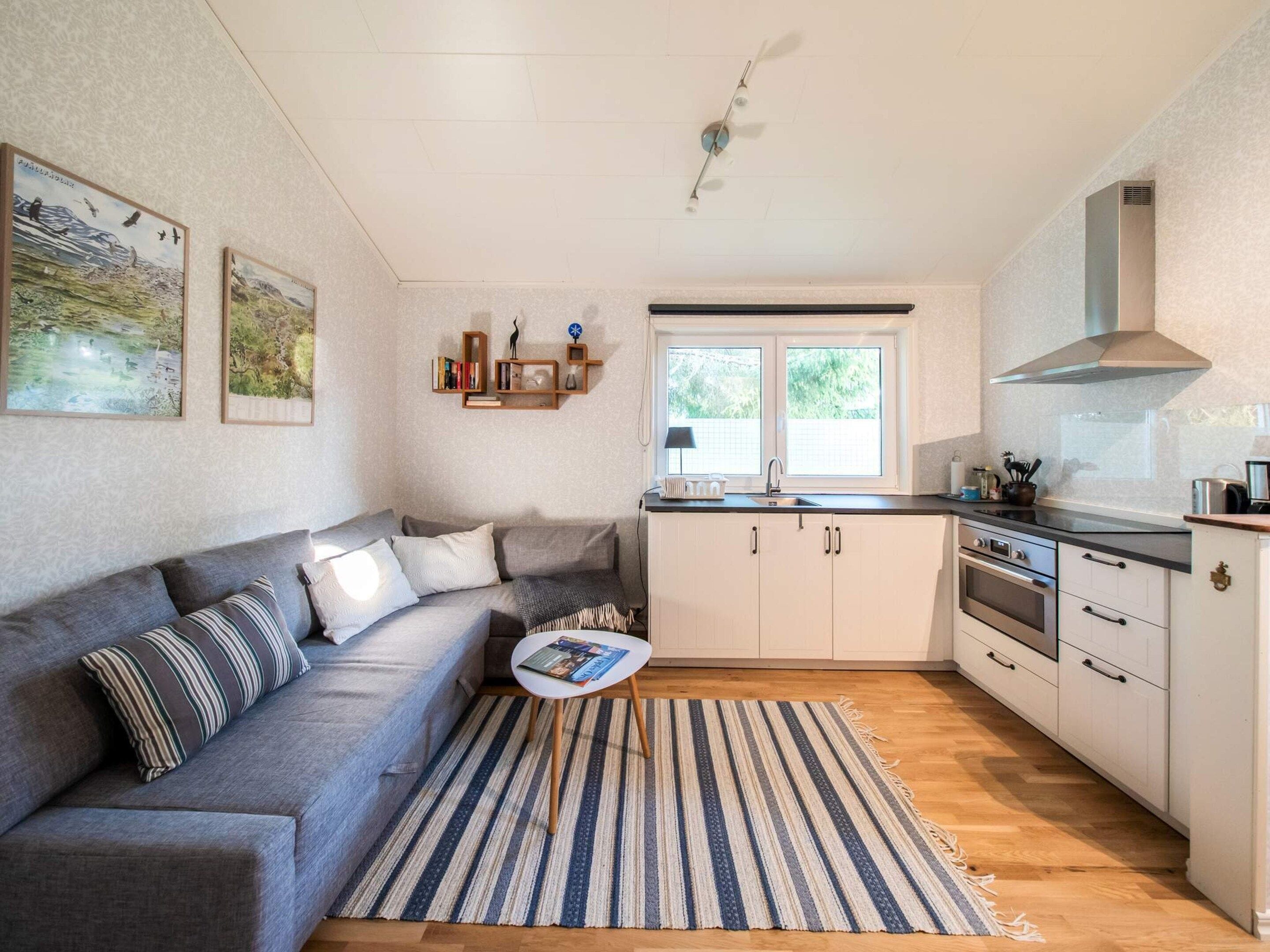 A compact living space with a sofa, a small coffee table, a kitchen with white cabinets, a stove, and a window with a view of greenery.