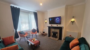 A living room with a fireplace, a television, and a dining area with chairs and a table. - Park Place 3 Bedroom House (North Northamptonshire)