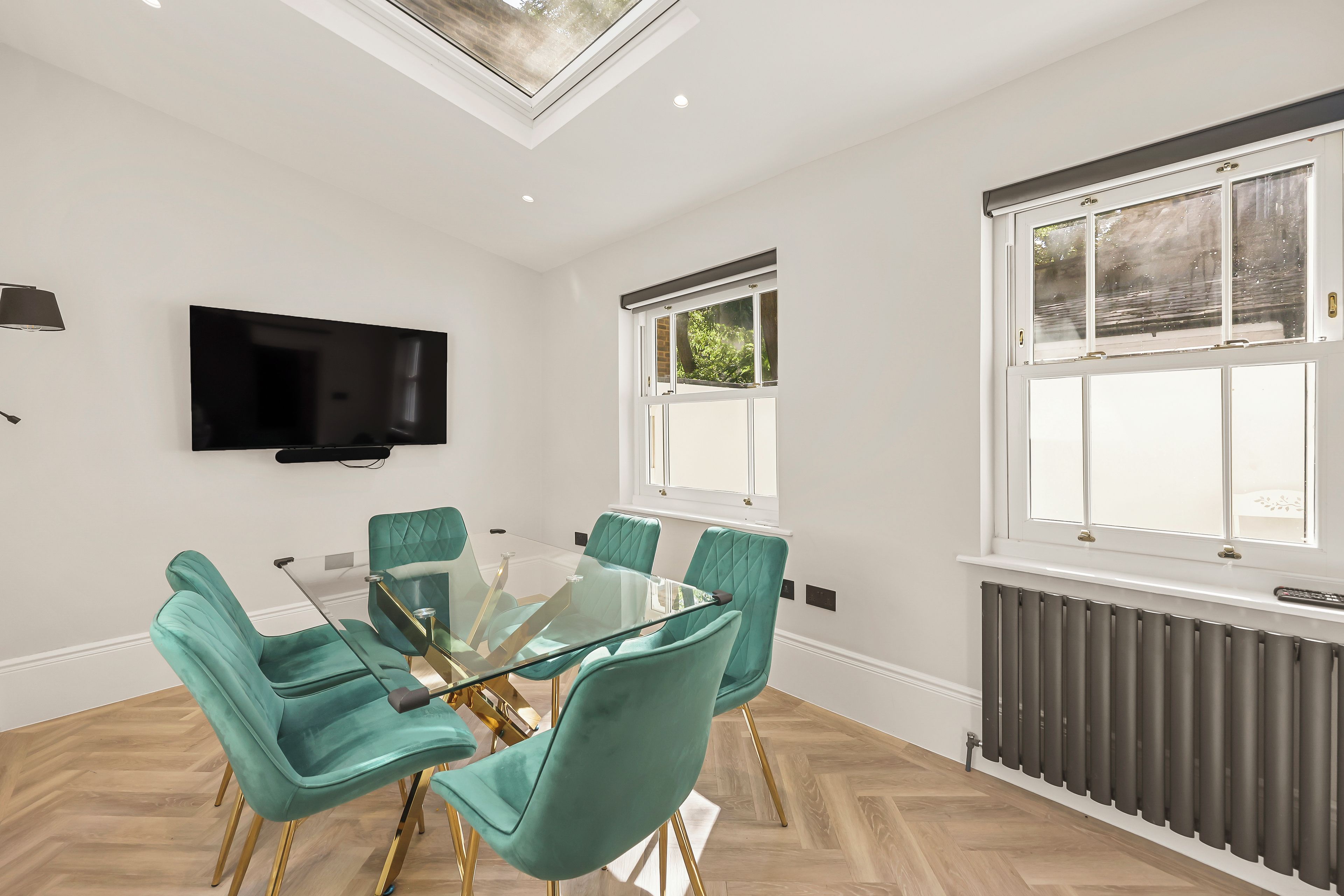 A modern dining area with a glass table and teal chairs, a flat-screen TV on the wall, and a window with a view of greenery.