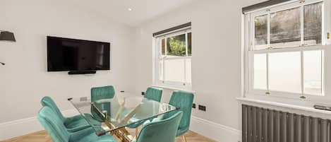 A modern dining area with a glass table and teal chairs, a flat-screen TV on the wall, and a window with a view of greenery.