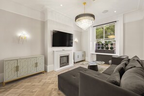 A modern living room with a flat-screen TV, a fireplace, a sofa, and a chandelier. - The Oakley Residence - Garden View Apartment (1) (London)