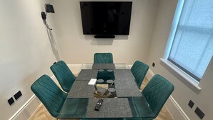 Et moderne spiseområde med et glasbord, turkise stole, et ophængt fjernsyn og et vindue med persienner.