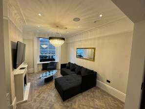 A modern living room with a television, a chandelier, a sofa, a coffee table, and a window with decorative plants. - The Oakley Residence - Garden View Apartment (1) (London)