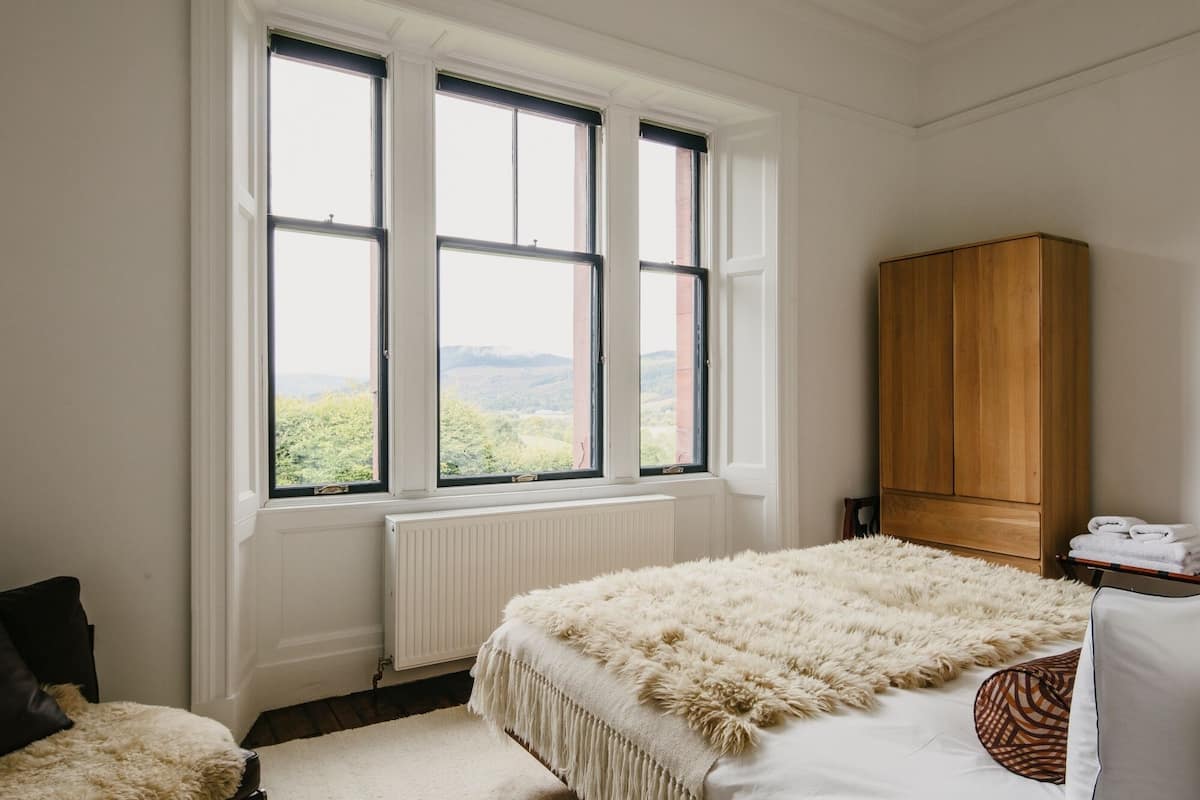 A bedroom with a large window, a wooden wardrobe, a bed with a fluffy blanket, and a radiator.
