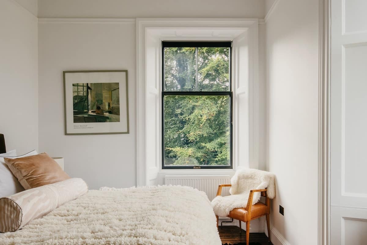 A bedroom with a large window, a framed painting, a bed with a fluffy white blanket, and a chair with a white fur throw.