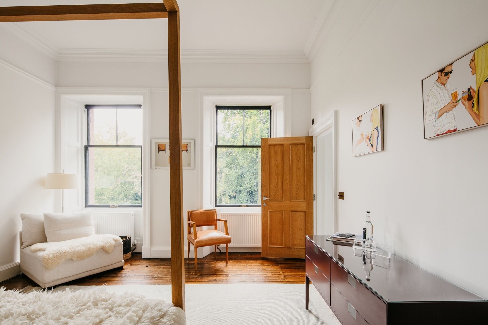 A room with a white sofa, a wooden chair, a bed with a white furry rug, a wooden door, and a dresser with a mirror.