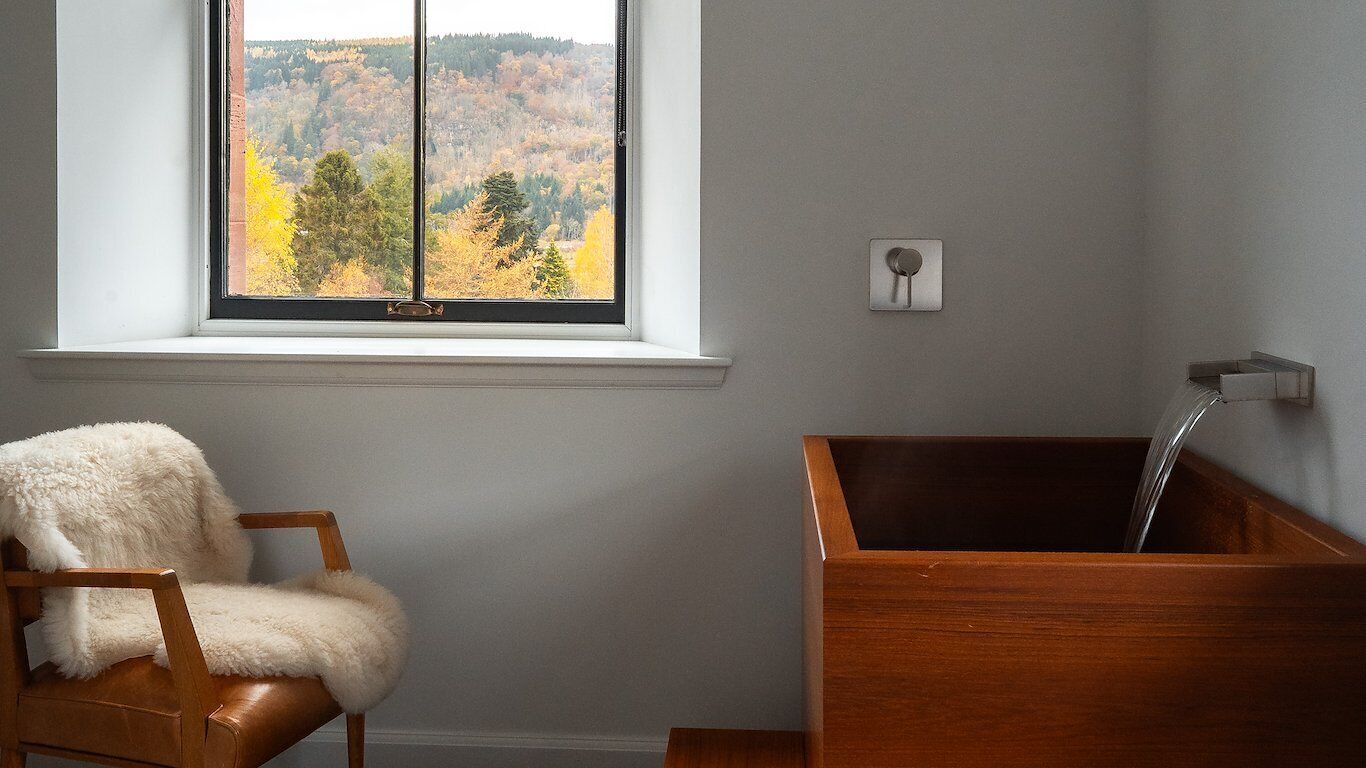 A bathroom with a wooden bathtub, a chair with a fluffy cover, and a window with a view of autumn trees.