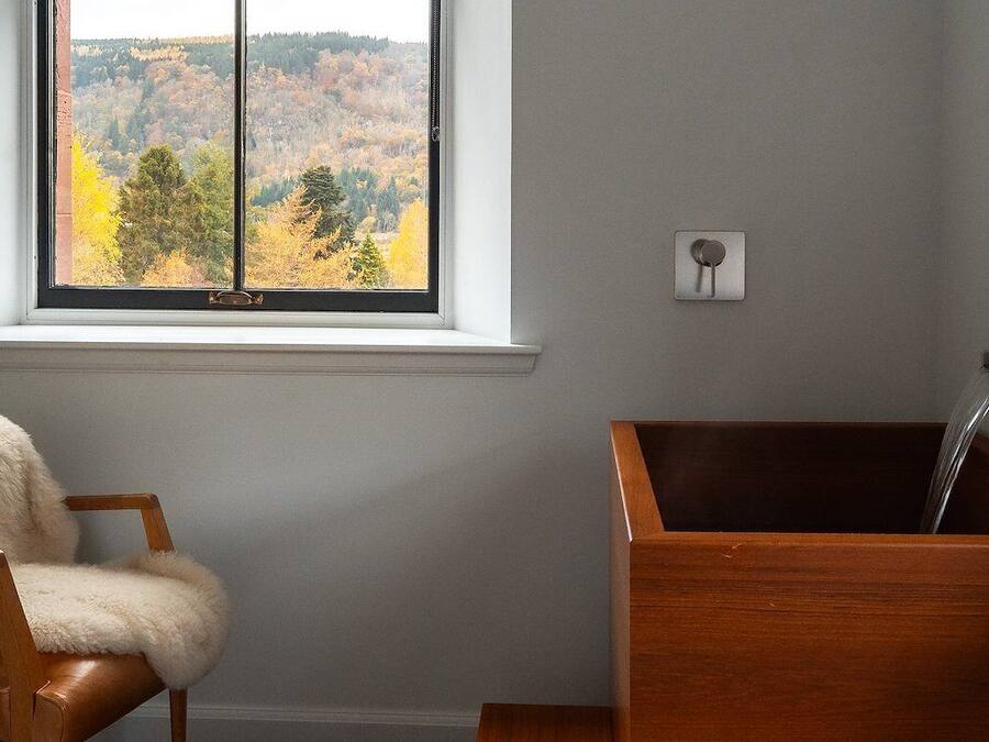 A bathroom with a wooden bathtub, a chair with a fluffy cover, and a window with a view of autumn trees.