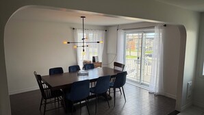 A dining area with a large table, six chairs, a modern chandelier, and a view through a glass door.