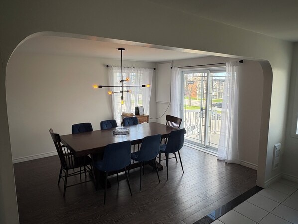 A dining area with a large table, six chairs, a modern chandelier, and a view through a glass door.