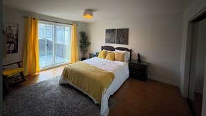 A bedroom with a bed, a nightstand, a potted plant, a window with curtains, and a door.