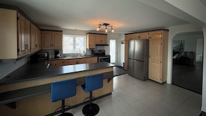 A modern kitchen with wooden cabinets, a stainless steel refrigerator, a large island with bar stools, and a view to the living area.