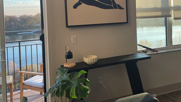 Una habitación con un ventanal grande, un cuadro abstracto enmarcado, un escritorio con una lámpara, un cuenco y una planta en maceta.