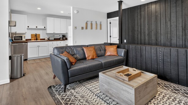 Un salon moderne avec un canapé en cuir foncé, une table basse en bois et une cuisine en arrière-plan.