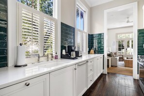 Une cuisine moderne avec des meubles blancs, une grande fenêtre équipée de stores, une machine à café et une vue sur l’espace de vie.
