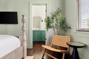 Un dormitorio con cama, una silla, una mesita, televisión y vista al baño.