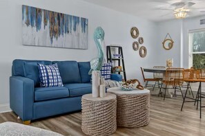 Ein Wohnzimmer mit blauem Sofa, zwei runden Flechtpoufs, einem Essbereich mit Holztisch und Stühlen sowie einem Deckenventilator.