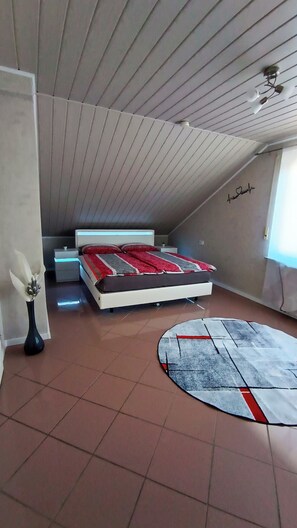Ein Schlafzimmer mit schrägverdeckter Decke, einem Bett mit roter und grauer Bettdecke, einem Nachttisch, einer Lampe und einem Teppich. Eine Vase mit Blumen steht ebenfalls dort.