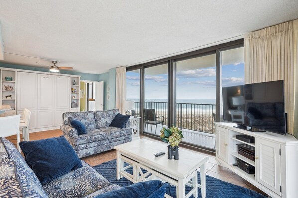 A spacious living room with a large TV, a sofa, a coffee table, and a balcony with a view of the beach. - 3 Bedroom, 2.5 Bath, Sea Colony Oceanfront Condo! (Bethany Beach)