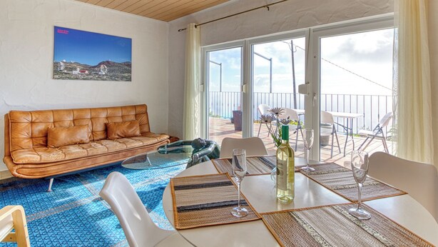 Una habitación con un sofá de cuero marrón, una mesa de centro de vidrio, una mesa de comedor con individuales y copas de vino, y vistas del exterior a través de grandes puertas corredizas de vidrio.