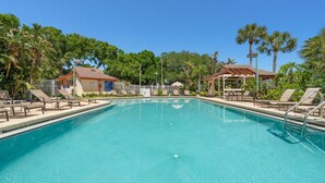 A clear swimming pool with lounge chairs and a covered seating area, surrounded by palm trees and a building with a sign. - Harbor Hideaway|Yard + Community Pool, Near Beach (Palm Harbor)