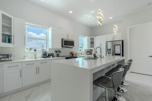 Een moderne keuken met witte kasten, een groot eiland met barkrukken, roestvrijstalen apparatuur en uitzicht op de omgeving.