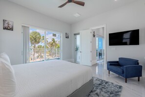 Een slaapkamer met een groot bed, een plafondventilator, een televisie en een balkon met uitzicht op palmbomen.