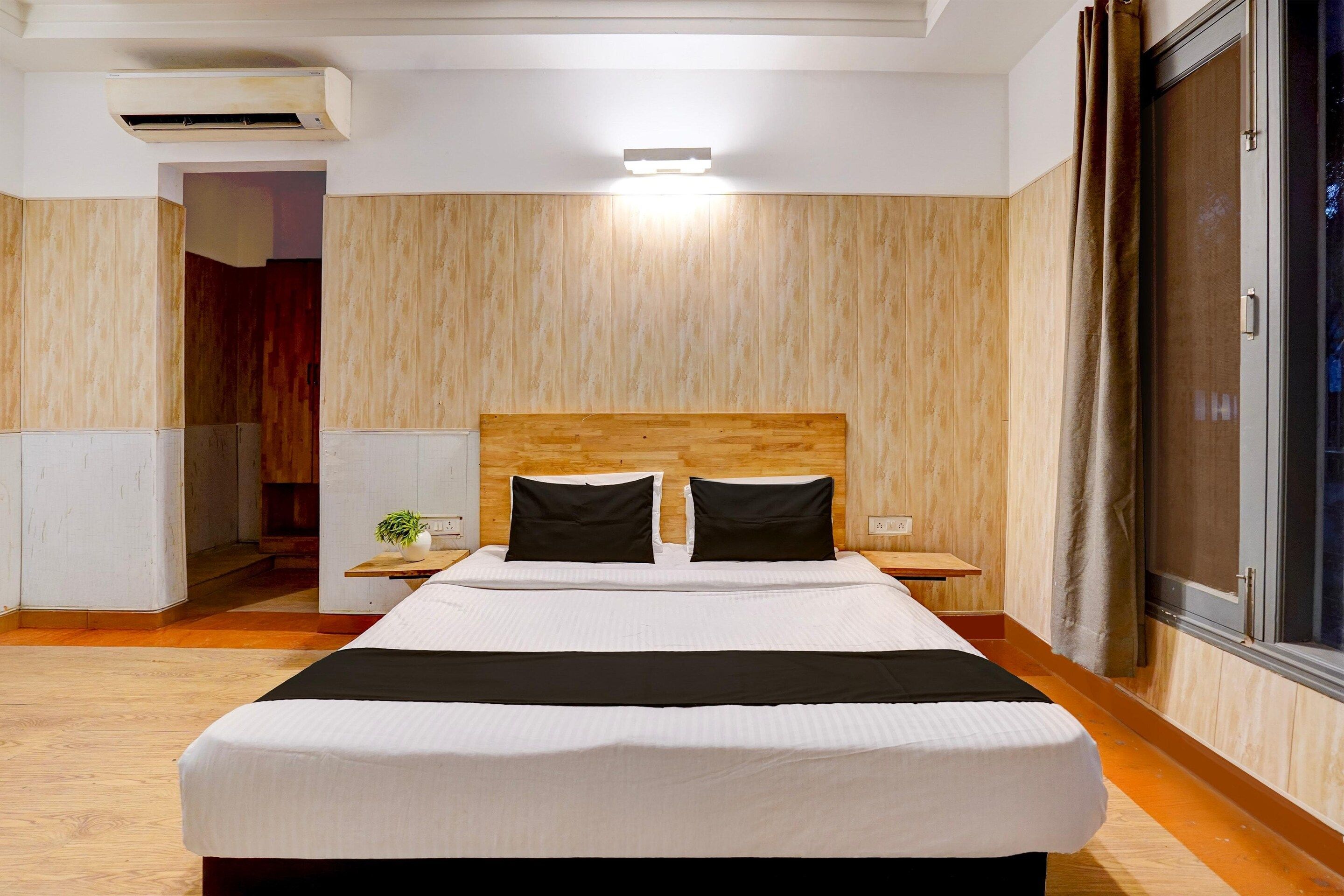 A hotel room with a wooden headboard, a large bed with white bedding, a nightstand, a bedside table, and a view of the outside.