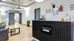 Een receptiebalie met de tekst 'TOWNHOUSE', en een bordje met 'Bedrijfsservice'. Er zijn plafondventilatoren, een klok en een wandplank met spullen.
