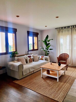 Un salon comprenant un canapé, une table basse, un fauteuil en osier, une grande plante et deux fenêtres avec des rideaux.