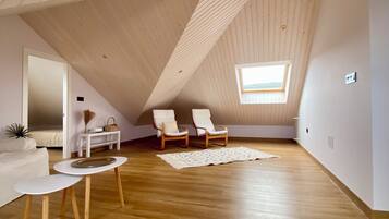 Un dormitorio en el ático con suelo de madera, un tragaluz, un sofá blanco, dos sillones, una mesita y una cesta decorativa.